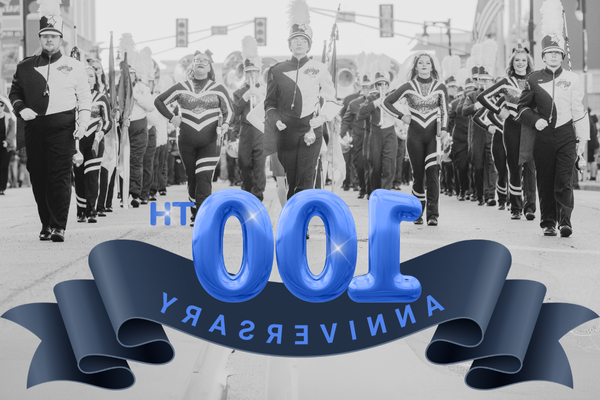 A black-and-white photograph of a marching band performing in a parade, with the text '100th Anniversary' in blue, bold letters overlaid on the image. The number '100' is styled as blue, balloon-like figures, with a blue ribbon beneath them containing the word 'Anniversary.' The band members are wearing traditional marching uniforms with plumes on their hats, and they are marching down a city street.